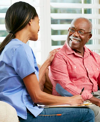 A caregiver in blue scrubs talks with a smiling older man in glasses